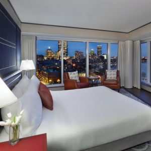 Modern luxury hotel suite with a large bed, two chairs, and floor-to-ceiling windows showcasing the city skyline at dusk.