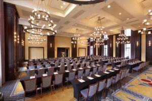 Elegant conference room with rows of tables and chairs, notepads and pens, large chandeliers, and tall windows letting in natural light.