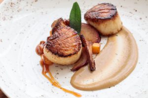 Two seared scallops on a white plate with brown sauce and roasted vegetables—a restaurant menu highlight, artfully arranged with herb garnish.
