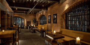 A dimly lit, cozy lounge in a historic Massachusetts hotel with brick walls, arched windows, and candlelit tables creates a warm, rustic atmosphere.