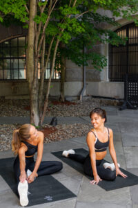 Two women in black sportswear stretching on yoga mats outdoors, smiling at each other under a tree with a building in the background.