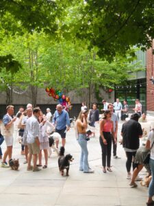 A group of people and dogs gather outdoors on a paved area, surrounded by trees and balloons, enjoying a social event.