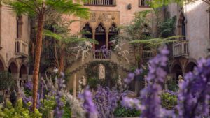 Lush courtyard garden with purple flowers, palm trees, statues, and a grand stone staircase in a historic building with arched windows and balconies.