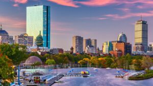 Boston city skyline at sunset with tall buildings, colorful sky, trees, and sailboats docked along the Charles River.
