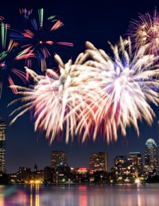 Colorful fireworks burst over Boston’s city skyline at night, reflecting on the water below with illuminated buildings and nearby accommodations in view.