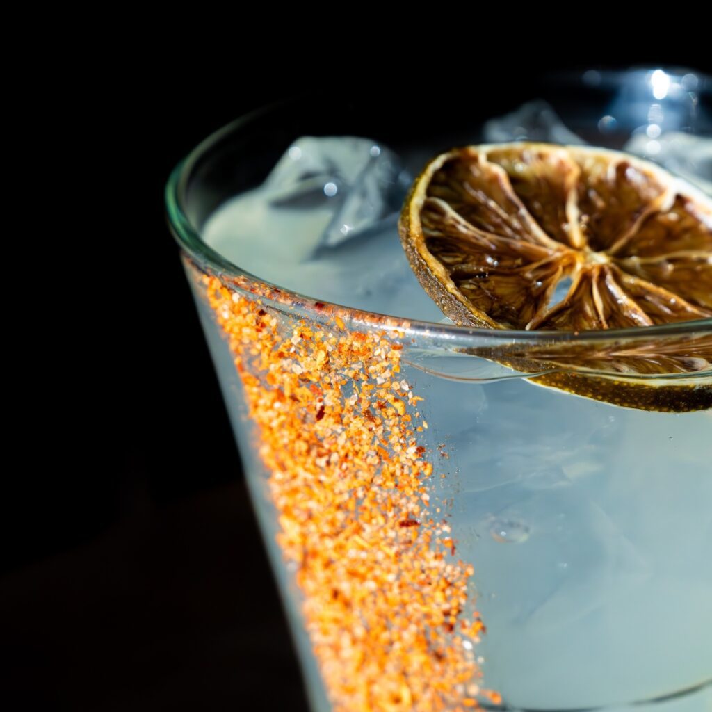 A close-up of a cocktail with ice, a dried lime slice, and chili salt rim in an American bar on Beacon Hill against a dark background.