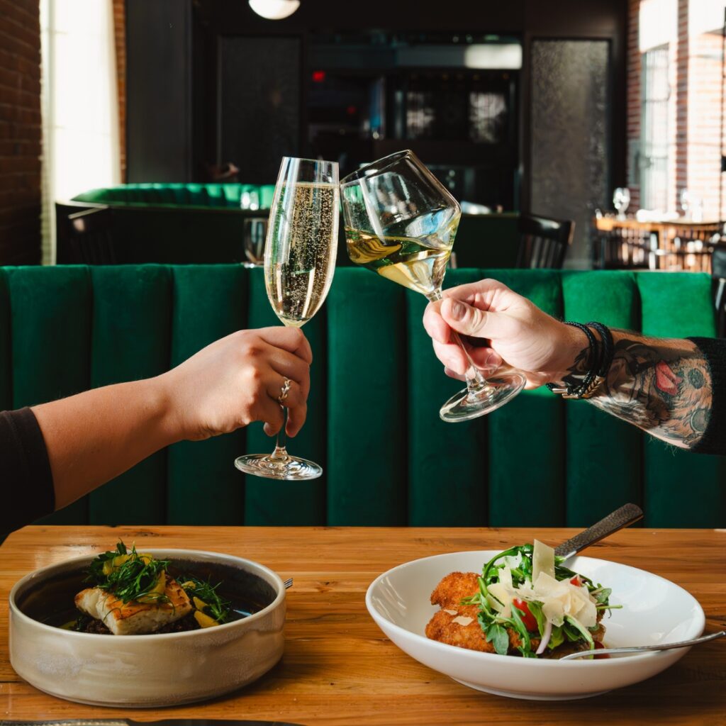 Two people clink champagne and wine glasses over gourmet dishes in a modern American restaurant with green booth seating in Boston’s Beacon Hill.
