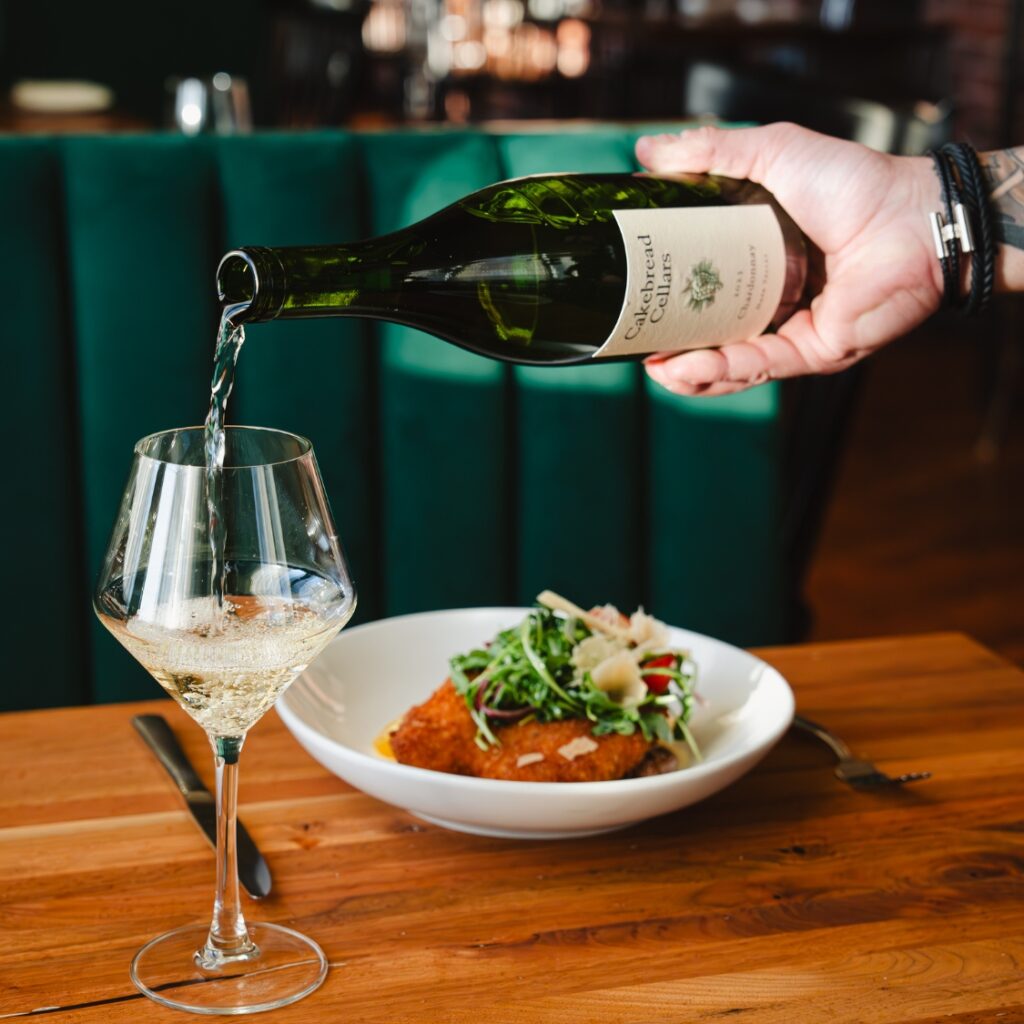 A hand pours white wine into a glass beside salad and breaded food on a wooden table in a Boston restaurant.