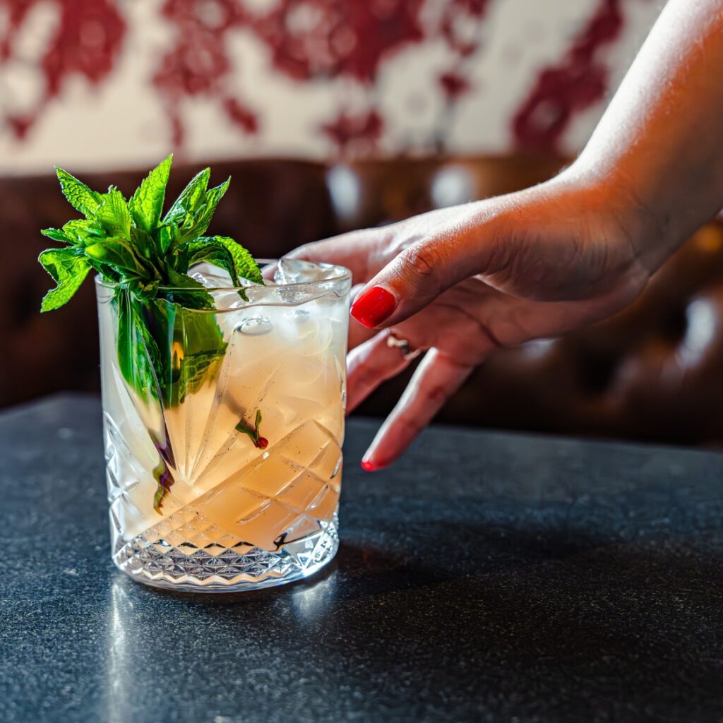 A hand with red nail polish reaches for a cocktail garnished with mint and ice, on a dark table at an American restaurant in Boston.