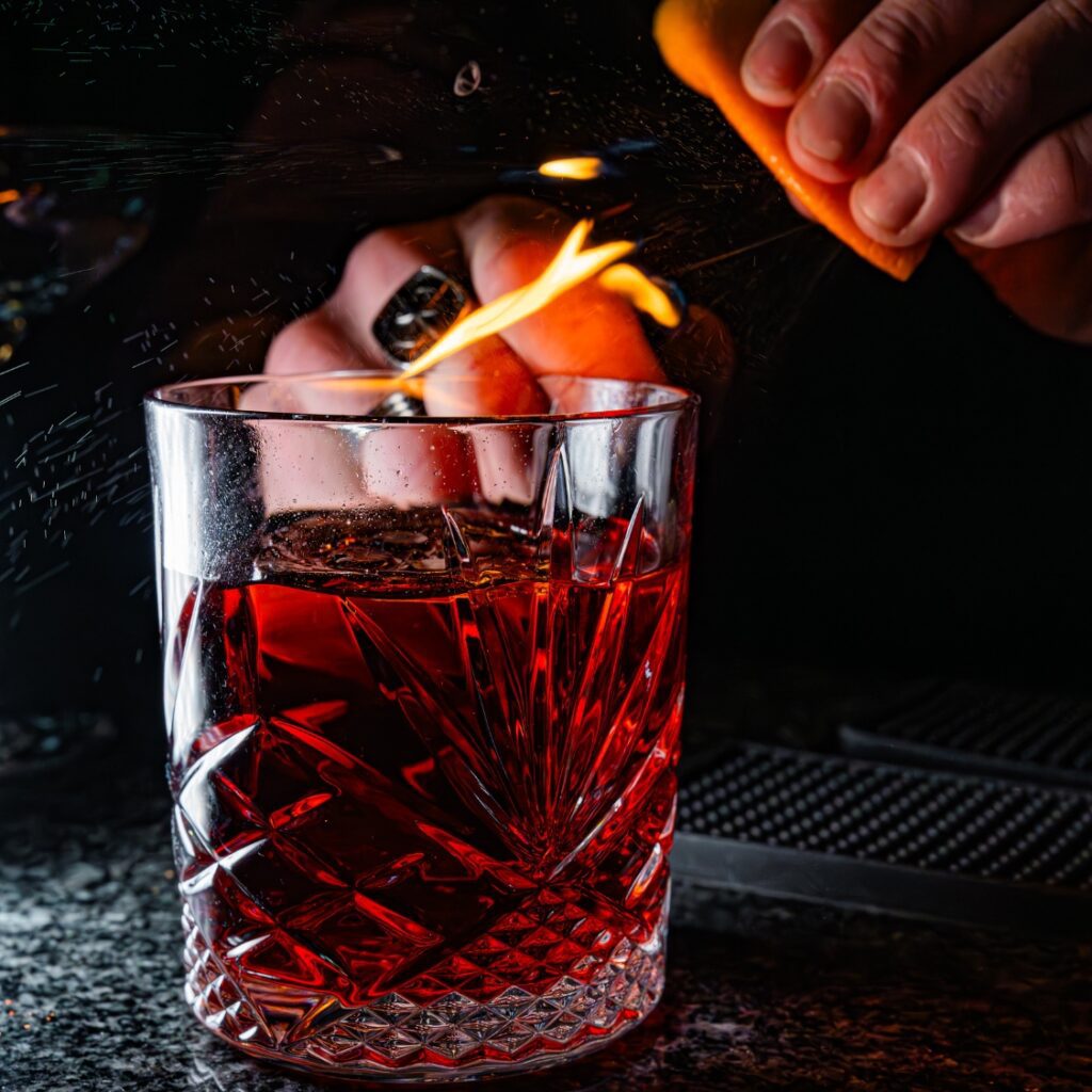 A hand squeezes an orange peel, igniting a flame over a red cocktail at a Boston bar counter with a dark Massachusetts background.