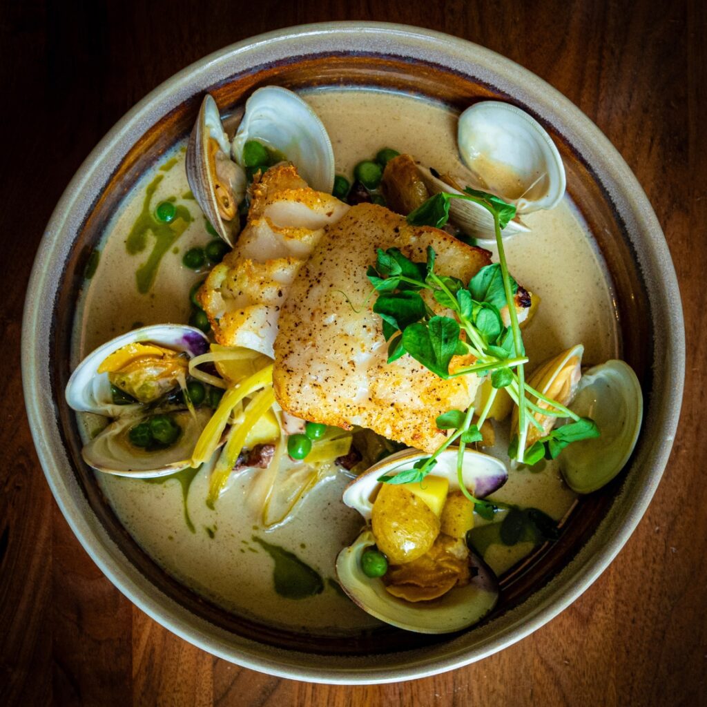 A bowl of seared fish with clams, potatoes, peas, and microgreens in a creamy sauce—classic American fare inspired by Boston, served on wood.