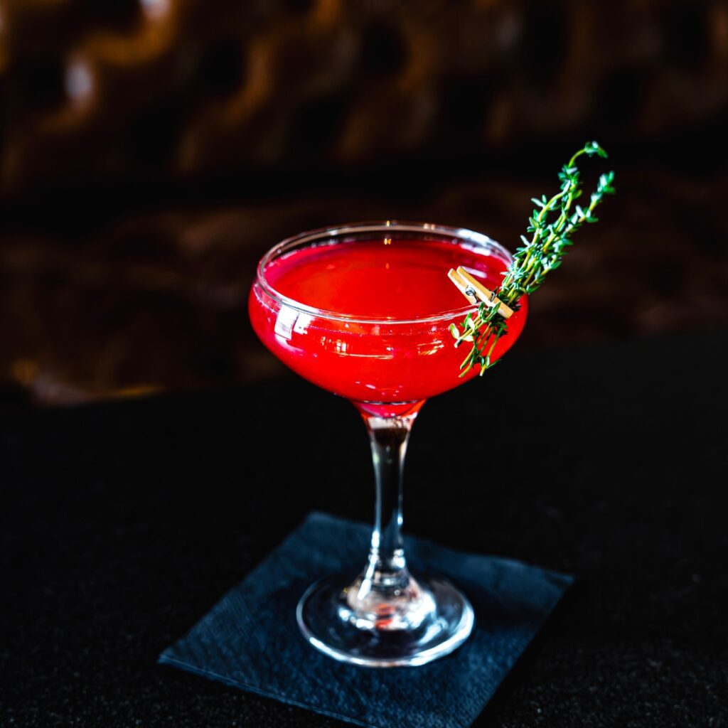 A vibrant red cocktail in a coupe glass, garnished with thyme, sits on a black napkin—elegant and perfect for a night out near Boston Common.