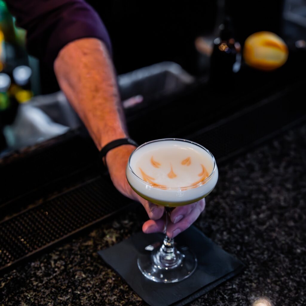 A bartender at a Beacon Hill restaurant serves a cocktail with a frothy top and orange garnish, placed on the bar counter with a black napkin underneath.