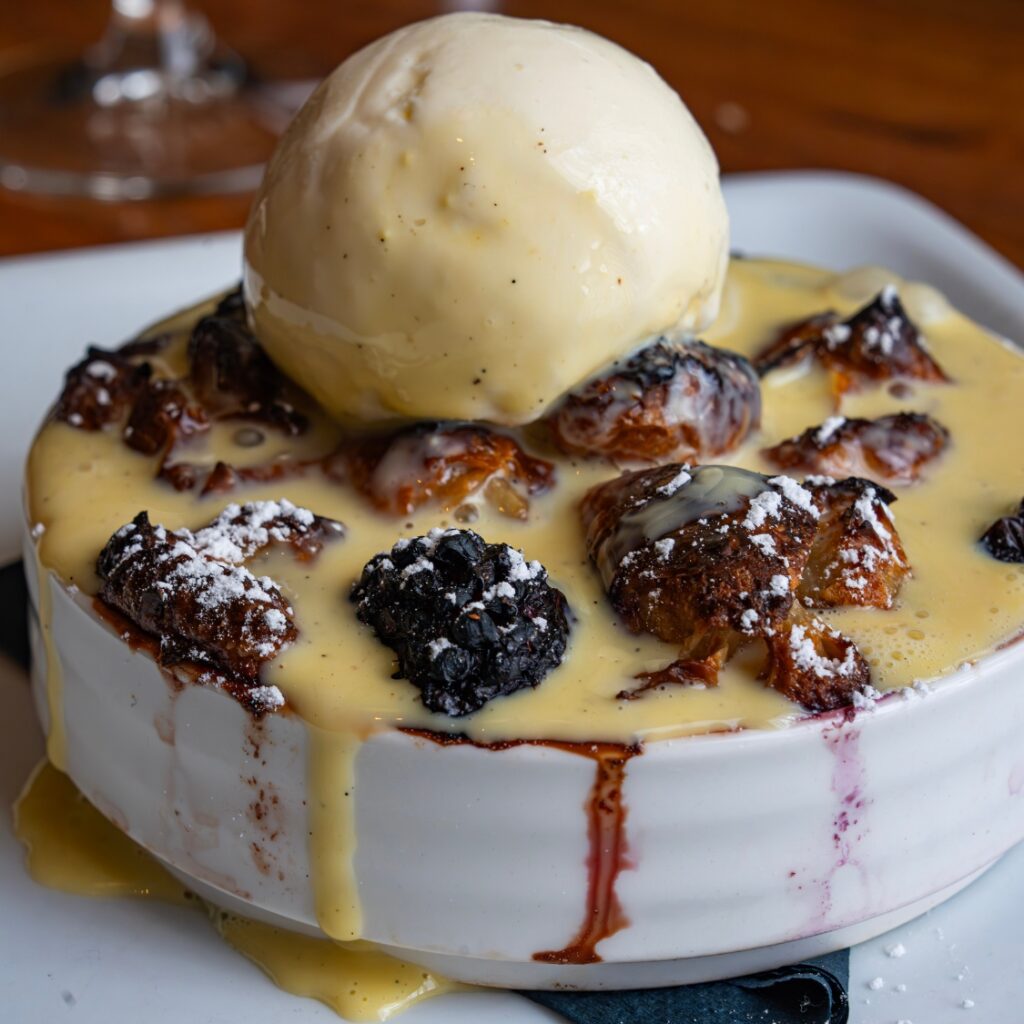 A warm bread pudding topped with blackberries, creamy sauce, and vanilla ice cream—served in a white dish at our Boston restaurant.