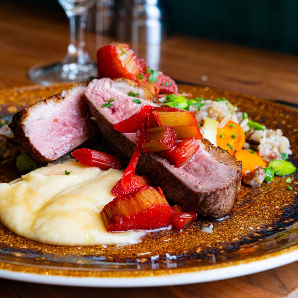 A plated dish features seared duck breast with rhubarb, creamy mashed potatoes, and grains on a brown ceramic plate near Boston Common.