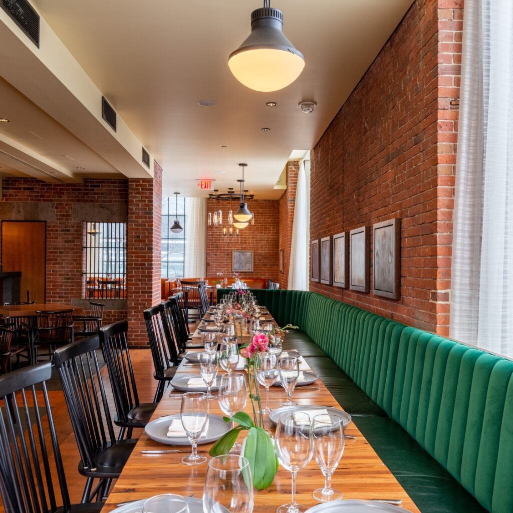 A long wooden table with green velvet benches and black chairs set for dining in a stylish American restaurant on Beacon Hill with brick walls.