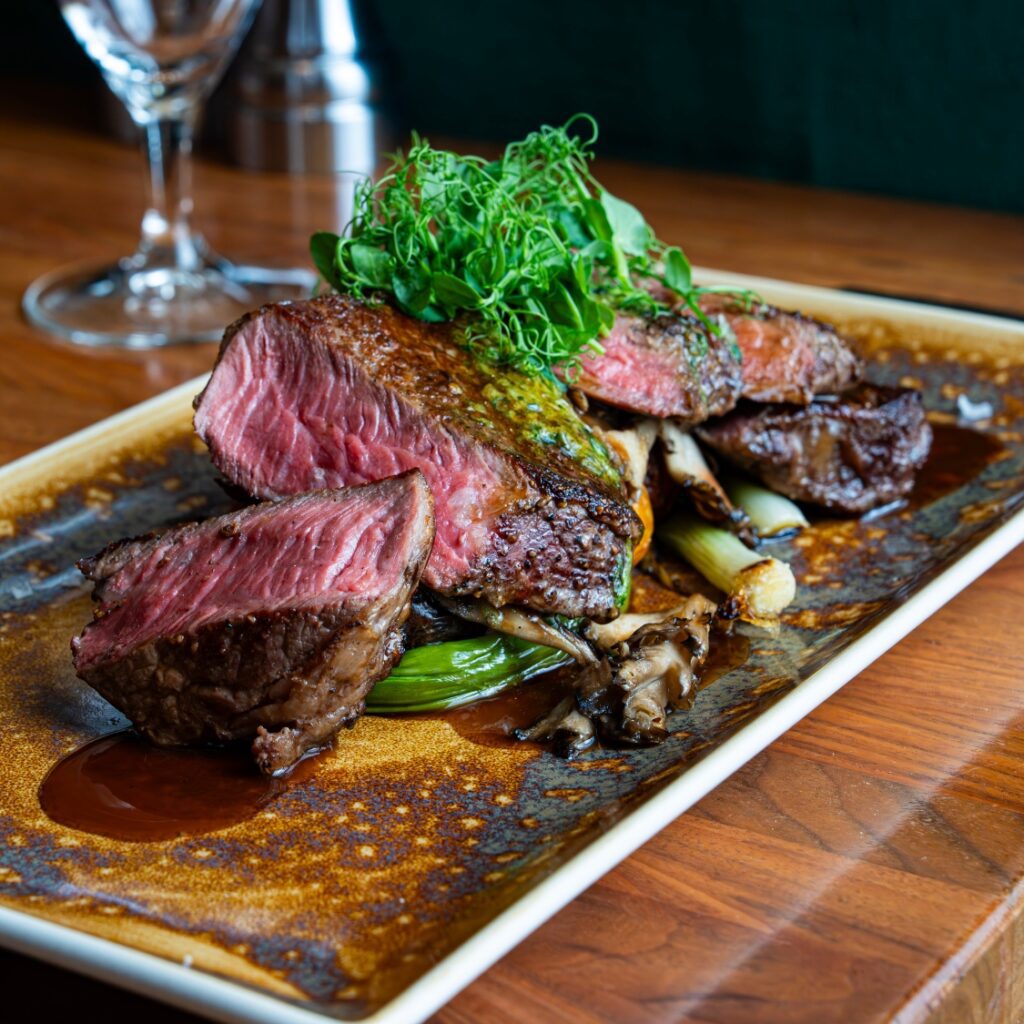 Sliced medium-rare steak topped with fresh greens, served with grilled vegetables and sauce at a Boston restaurant on a rectangular plate.