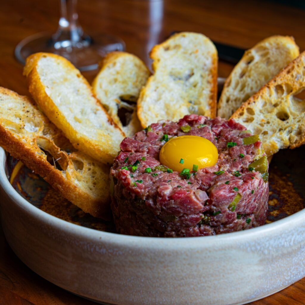 A plate of steak tartare topped with a raw egg yolk, garnished with chives, served with toasted baguette slices near Boston Common.