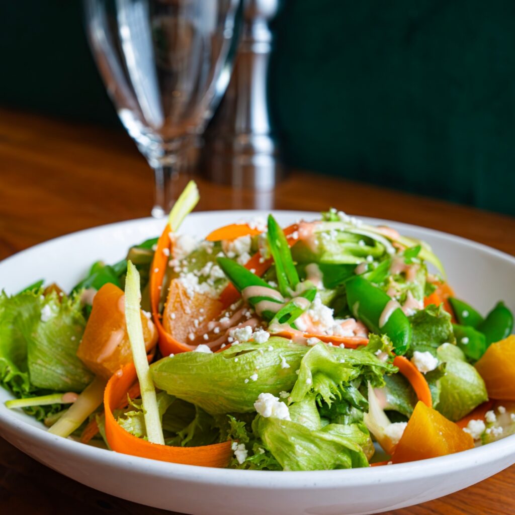 A fresh salad with lettuce, carrot ribbons, snap peas, and yellow beets served in a white bowl on a wooden table in Beacon Hill, Boston.