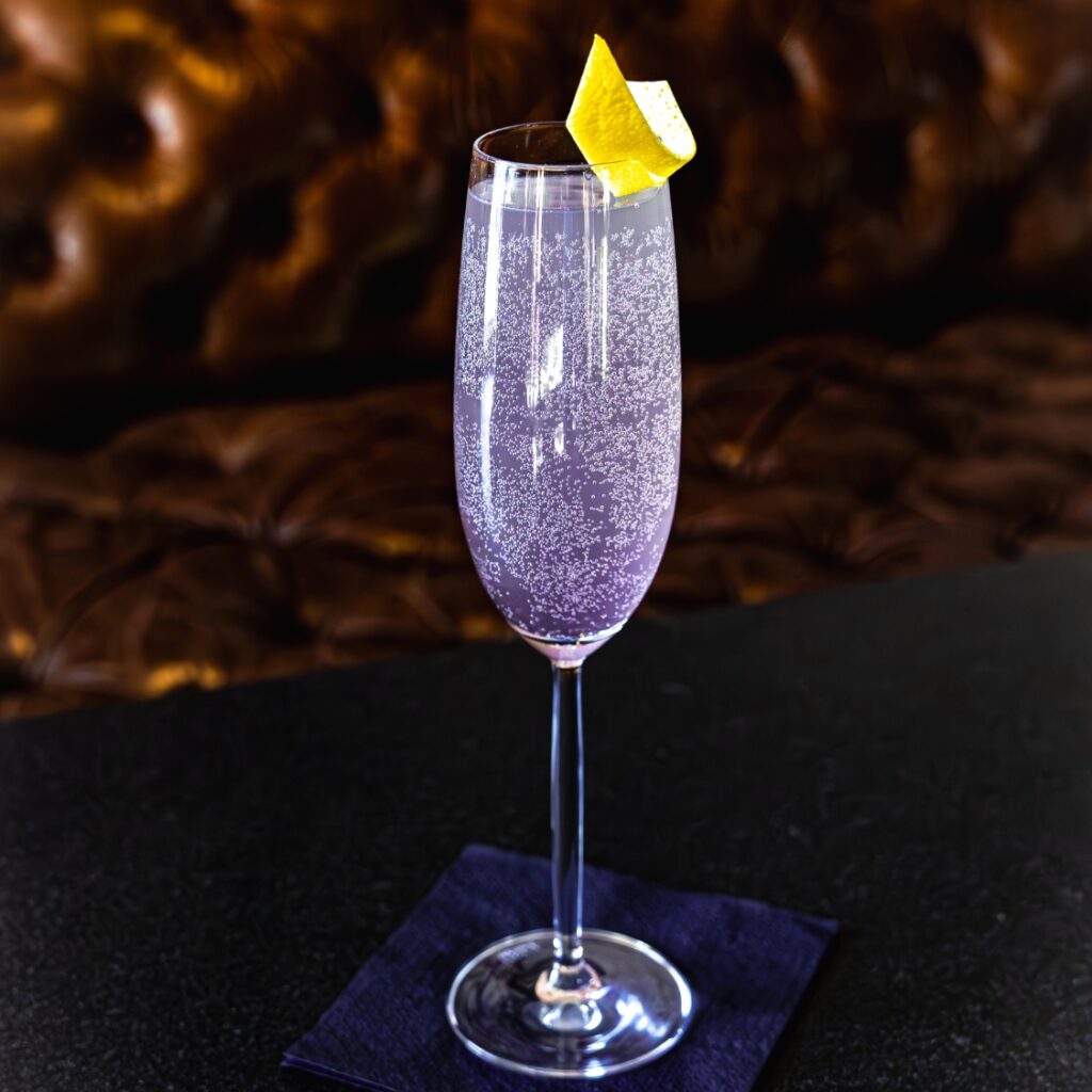 A sparkling lavender cocktail in a Boston restaurant, served in a flute with a lemon twist, set on a dark napkin and table with a brown tufted background.