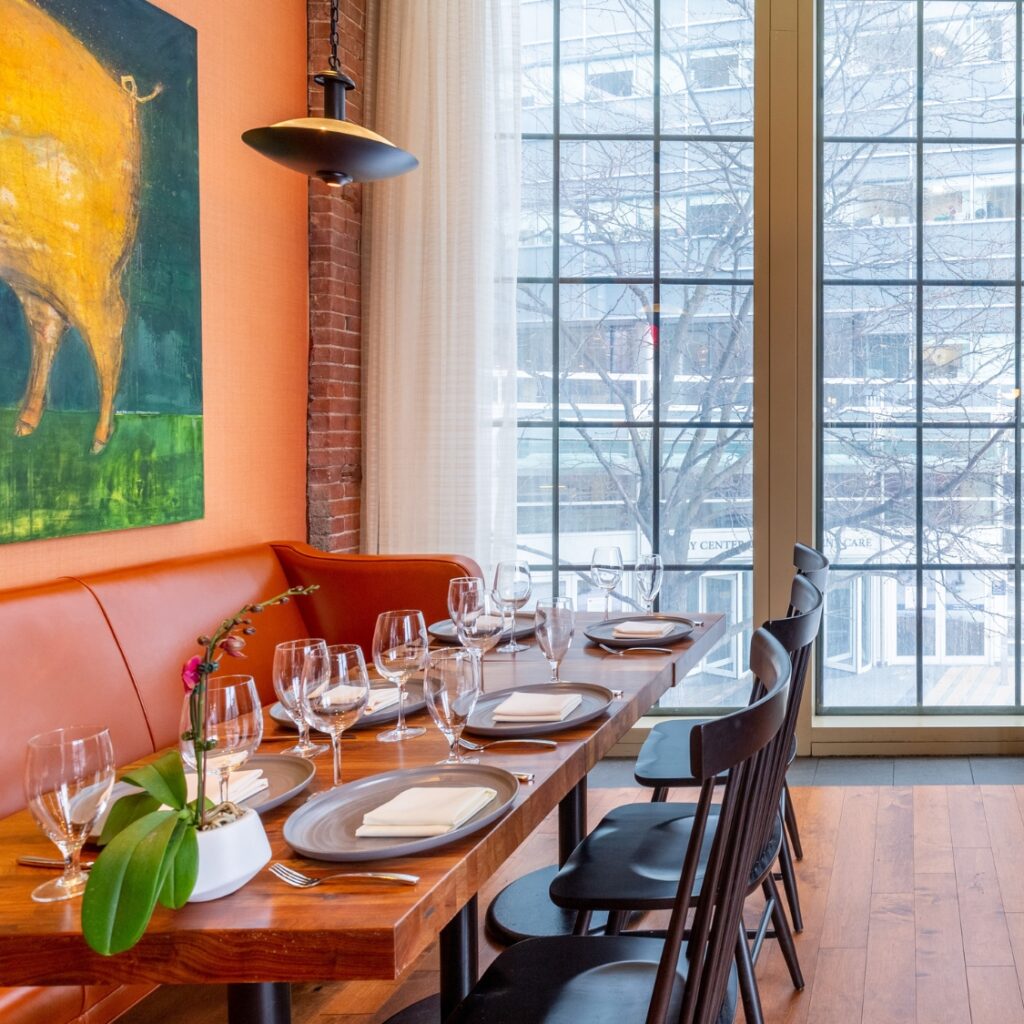 A modern Boston restaurant table set for eight with wine glasses, next to large windows and an orange wall featuring a pig painting.