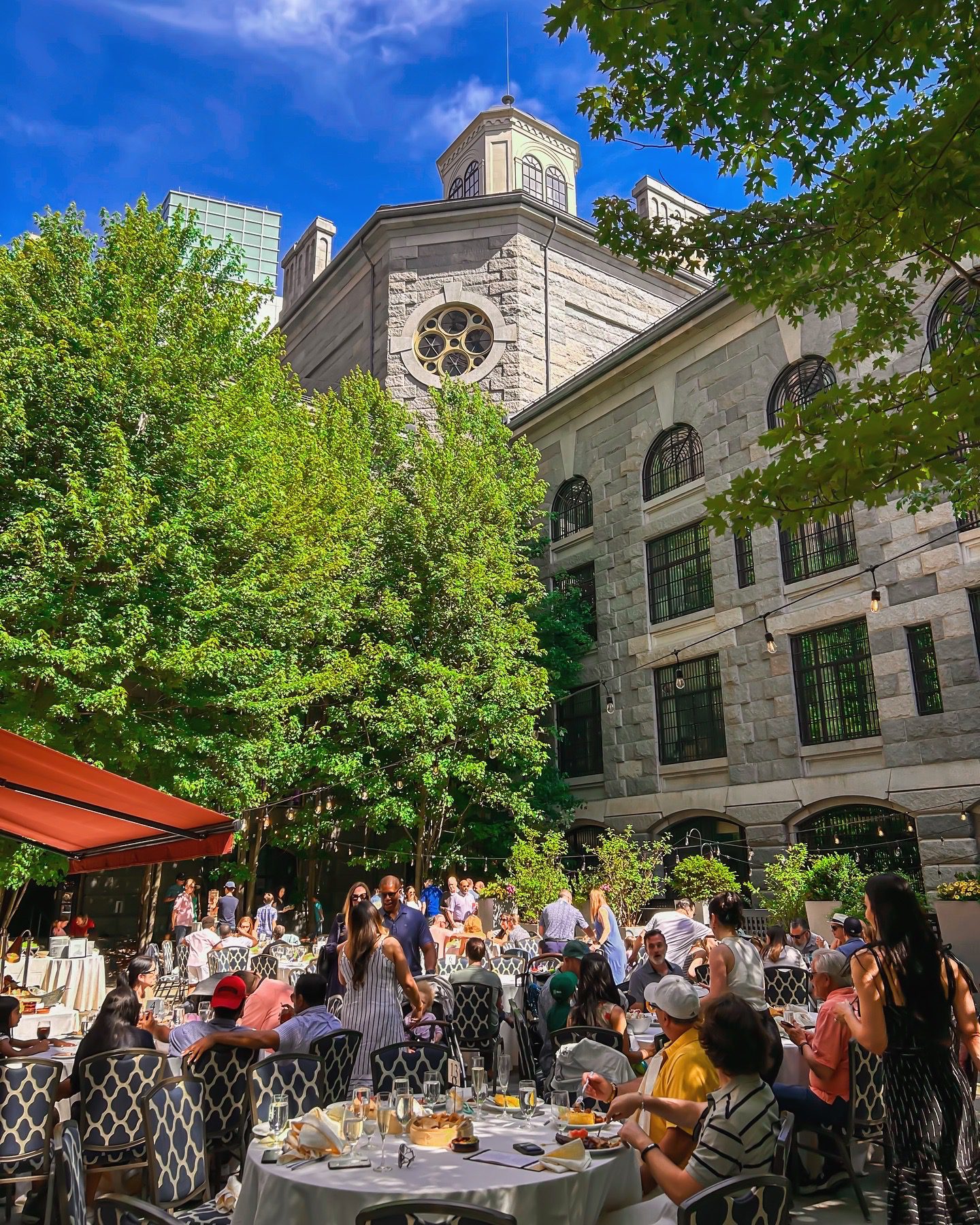 Father's Day People dine outdoors at tables in a lush garden patio beside a historic stone hotel on a sunny day.