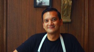 A smiling man in a black shirt and white apron sits in front of a wooden wall with framed art and decor in the background.