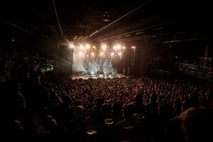 A large crowd watches a band perform on a brightly lit stage in a historic Boston, MA indoor concert venue.