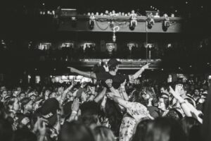 A crowd surfer is lifted by concertgoers at a packed historic Boston music venue, with people cheering and reaching up in a lively atmosphere.