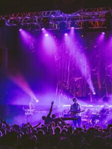 A band performs on stage in historic Boston, MA, under purple lights with a cheering crowd and dramatic draped backdrop at a concert.