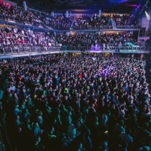 A large crowd fills an indoor concert venue in Boston, MA, with multiple balconies and colorful stage lights illuminating the audience.