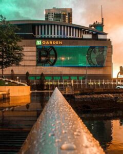 Sunset view of TD Garden in Boston, MA, with Bruins and Celtics logos on the arena and nearby hotel lights reflecting on the water.