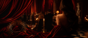 A woman in an elegant red gown faces a candlelit, crowded ballroom. A golden crown and glass of champagne rest on red velvet nearby.