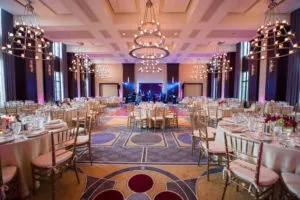 Elegant banquet hall with round tables, gold chairs, chandeliers, and a stage set for an event; purple lighting accents the spacious room.