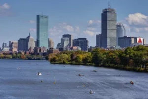 Rowers on the Charles River with the Boston skyline and skyscrapers in the background on a sunny day.