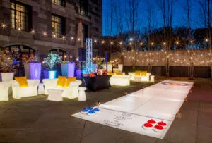 Outdoor lounge in Boston with glowing furniture, string lights, a curling game, and central cocktail bar at The Yard creates a festive atmosphere at dusk.