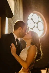 A bride and groom share a kiss in warm sunlight, standing in front of a round stained glass window inside a church.