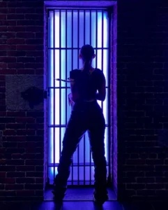 A person stands silhouetted before vertical bars and a bright blue light, evoking a moody lounge vibe amidst dark brick walls.