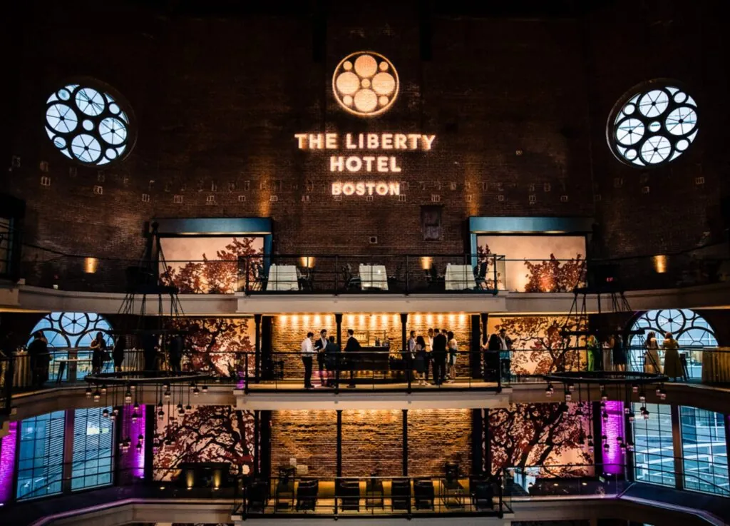 Lobby of The Liberty Hotel in Boston's luxury Beacon Hill, featuring large round windows, illuminated signage, and guests mingling on balconies.