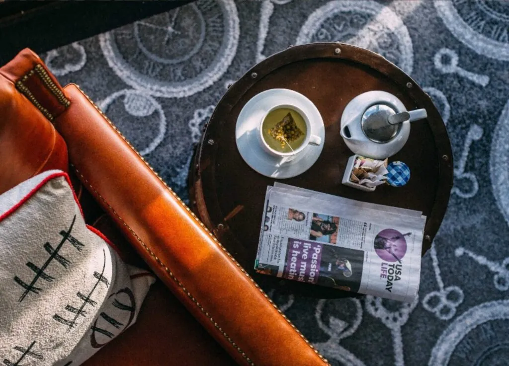 A cup of tea, a teapot, and newspaper await on a round table in a luxury Beacon Hill hotel beside a brown leather chair with a patterned pillow.
