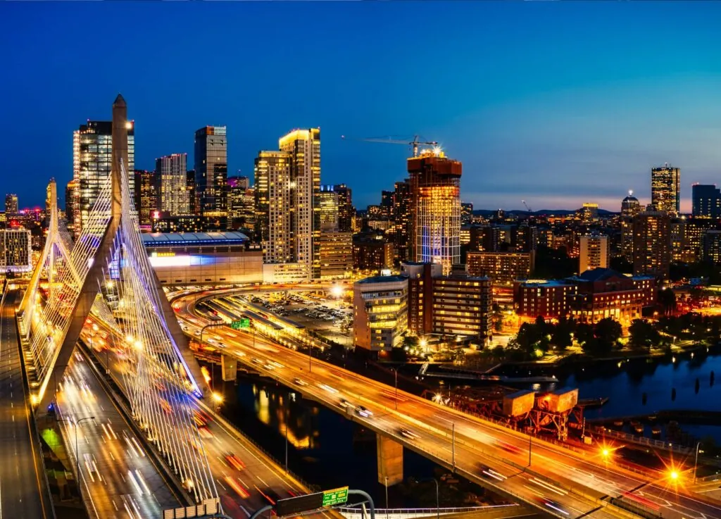A luxury photo of a brightly lit cityscape at dusk, featuring tall buildings, a cable-stayed bridge, and glowing highways over a river.