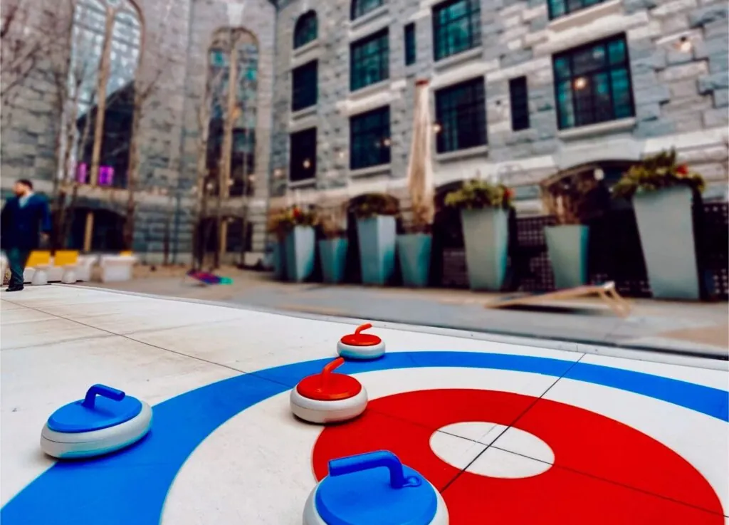 Outdoor curling game with red and blue stones on a target in a city plaza; photo features tall buildings and large planters in the background.