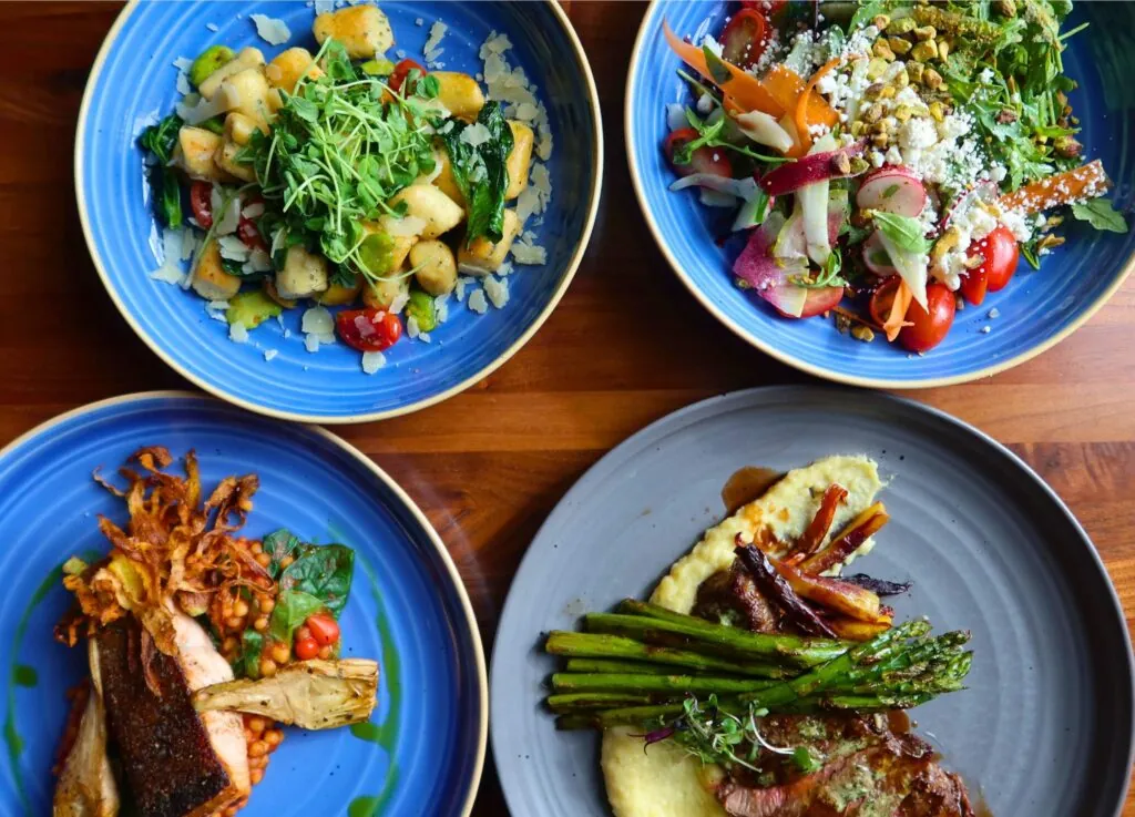 A luxury photo of four colorful plated dishes—gnocchi, salad, salmon, and steak—artfully arranged on a wooden table in a hotel dining setting.