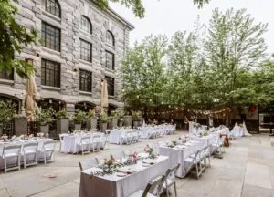 Luxury outdoor event space with white-clothed tables, string lights, and trees beside a stone building—photo-perfect for celebrations or weddings.