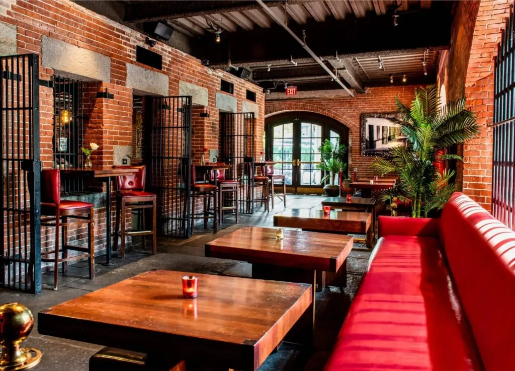 A cozy bar with luxury touches, exposed brick, red seating, wooden tables, tall chairs, potted plants, and gallery-style warm lighting.