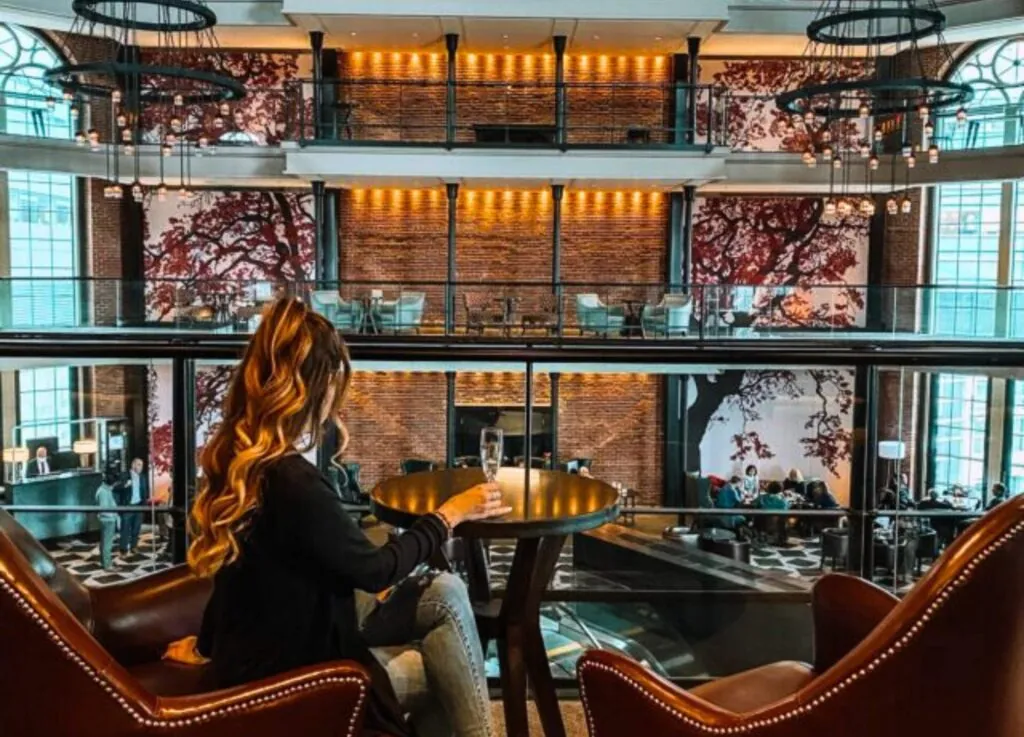 Woman with long wavy hair sits at a table with a drink in a stylish hotel lounge, overlooking Beacon Hill through large windows and intricate wall art.