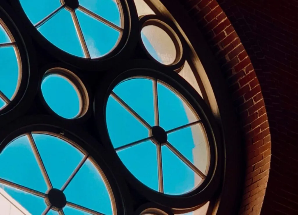 Luxury circular stained-glass window with geometric patterns set in a brick wall, perfect for a hotel photo with bright blue sky beyond the glass panes.