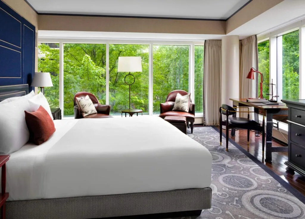 Bright luxury hotel room with a large bed, leather chairs, desk, and windows overlooking green trees near Beacon Hill.