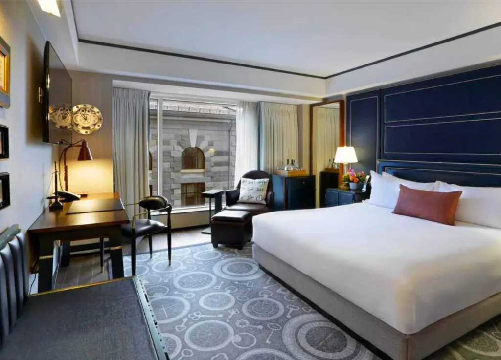 A luxury hotel room with a large bed, desk, armchair, TV, and window offering a stunning photo-worthy view of a stone building courtyard.