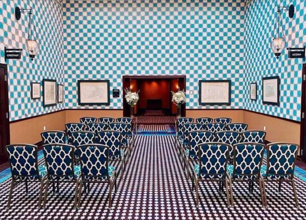 A small luxury event room on Beacon Hill features blue patterned walls, floral arrangements, framed photos, and rows of elegant chairs.